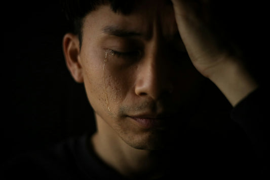 Asian man with tear rolling down side of face alone in dark room as he suffers suicidal ideations and thoughts of suicide